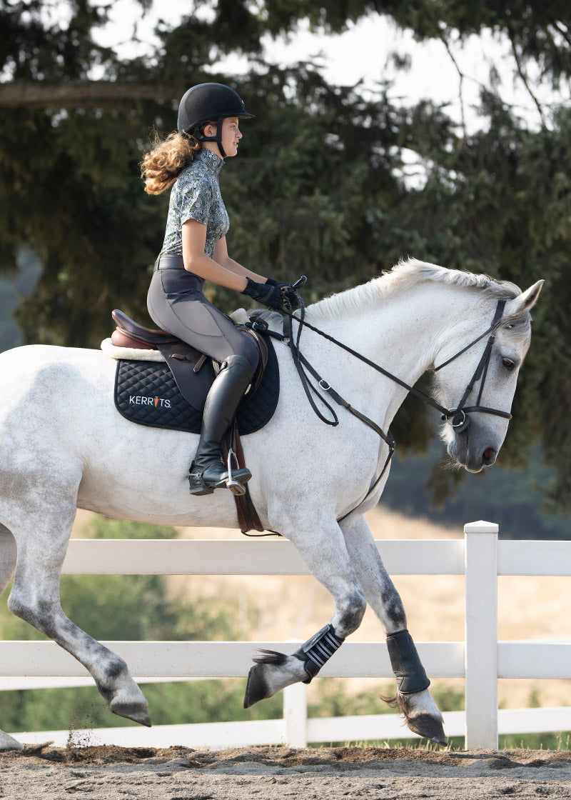 Equestrian riding a horse while wearing the Coolcore® Full Leg Tech Tight in Cinder, showcasing the full-leg silicone grip in action.