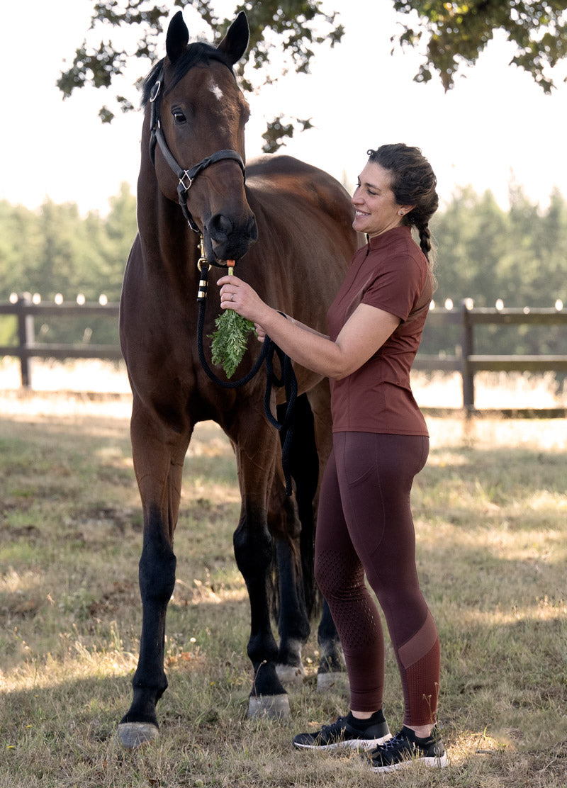 Rider wearing the Free Style Knee Patch Pocket Tight while feeding a horse a carrot, illustrating practical use in a barn setting.