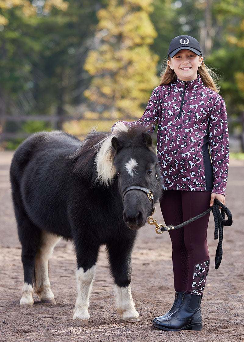 Full-body view of a young equestrian standing in a sunny outdoor setting, wearing the Kerrits Kids Thermo Tech™ Full Leg Tight and a matching riding top