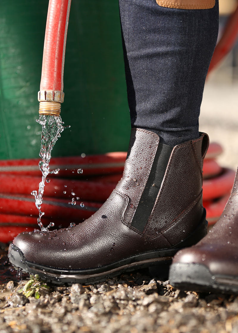 Individual with water hose next to Woodstock Waterproof Barn Boot in dark bay, illustrating waterproof feature and non-slip traction