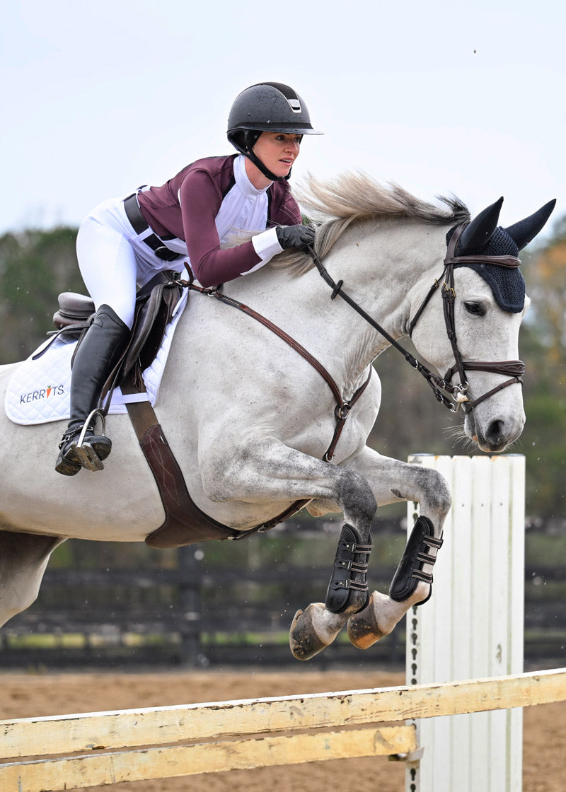 Rider wearing the Affinity® Pro Knee Patch Breech in white, jumping her horse in an arena.